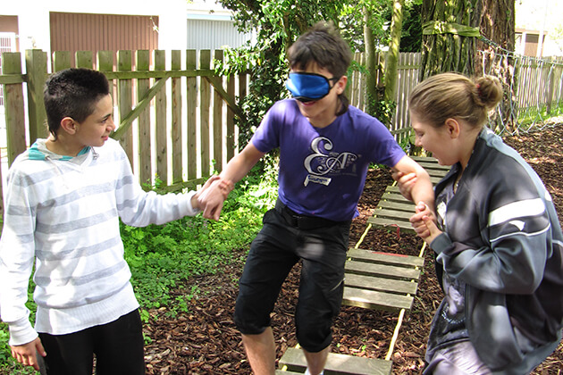 Zwei Personen helfen einer weiteren Person mit verbundenen Augen über einen Kletterpfad zu laufen Zwei Personen helfen einer weiteren Person mit verbundenen Augen über einen Kletterpfad zu laufen