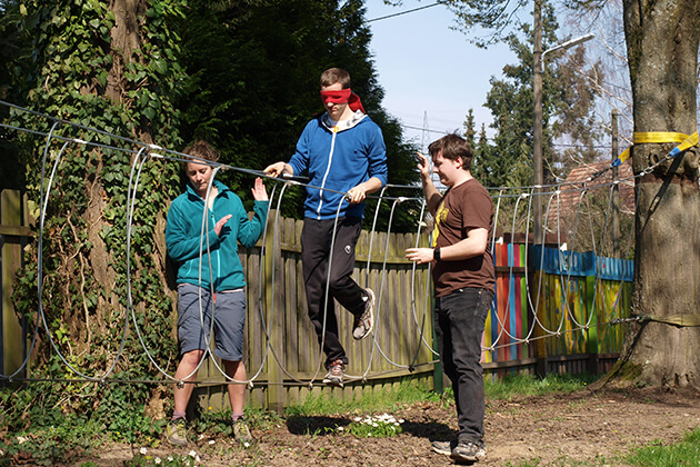 Eine Person läuft mit verbundenen Augen über ein Balanceseil. Zwei Personen helfen dabei. Eine Person läuft mit verbundenen Augen über ein Balanceseil. Zwei Personen helfen dabei.